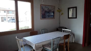 A dining area with a table set for four, chairs, a window with a view of a building and parked car, and wall decorations including a framed picture and hanging ornaments.