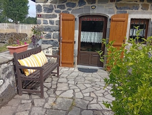 Un bâtiment en pierre avec un banc en bois, une plante en pot et une porte ornée d’un rideau en dentelle.