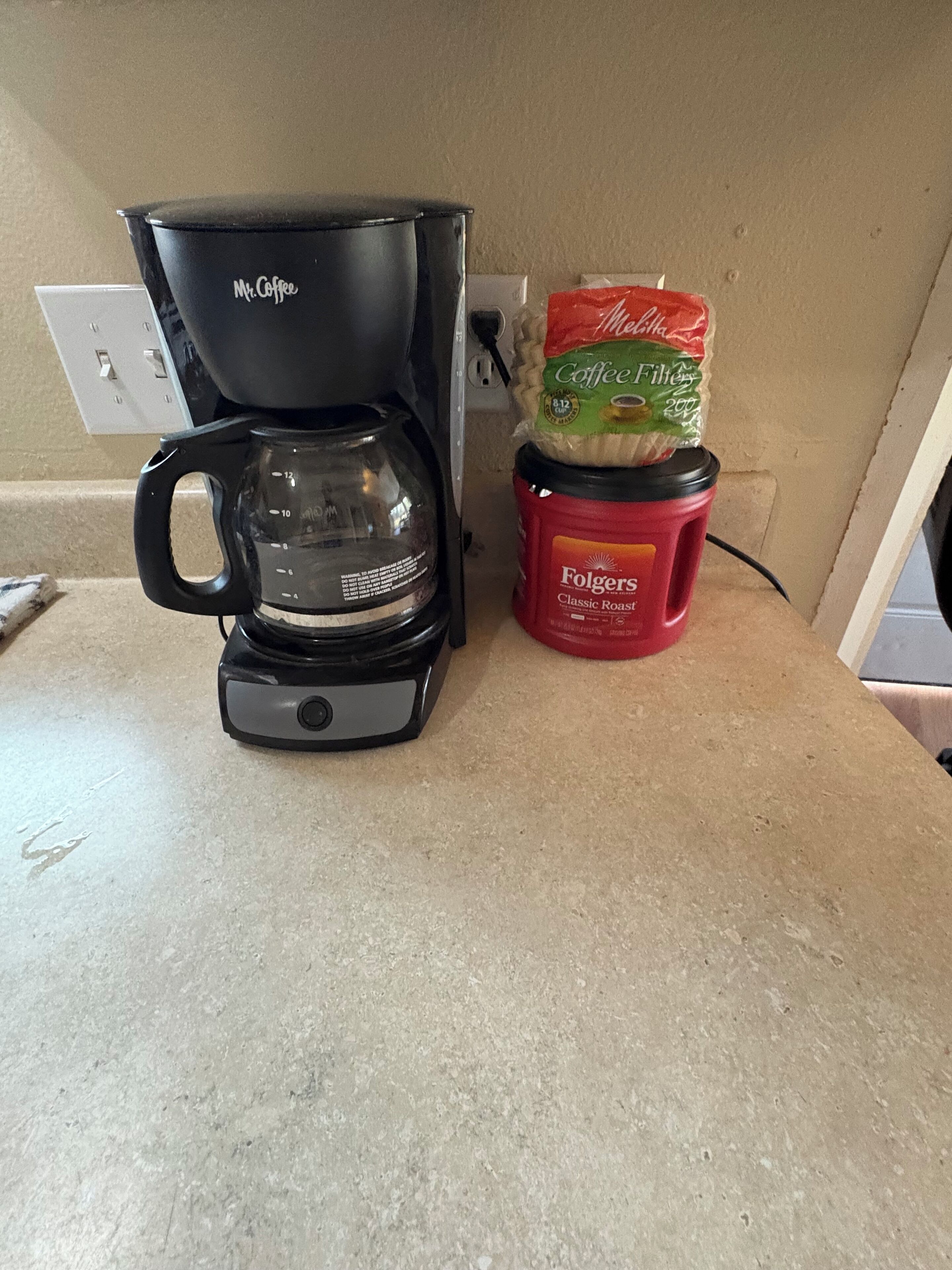 Una cafetera con jarra de vidrio, una bolsa de café Folgers y un contenedor rojo de Folgers sobre una encimera de cocina.