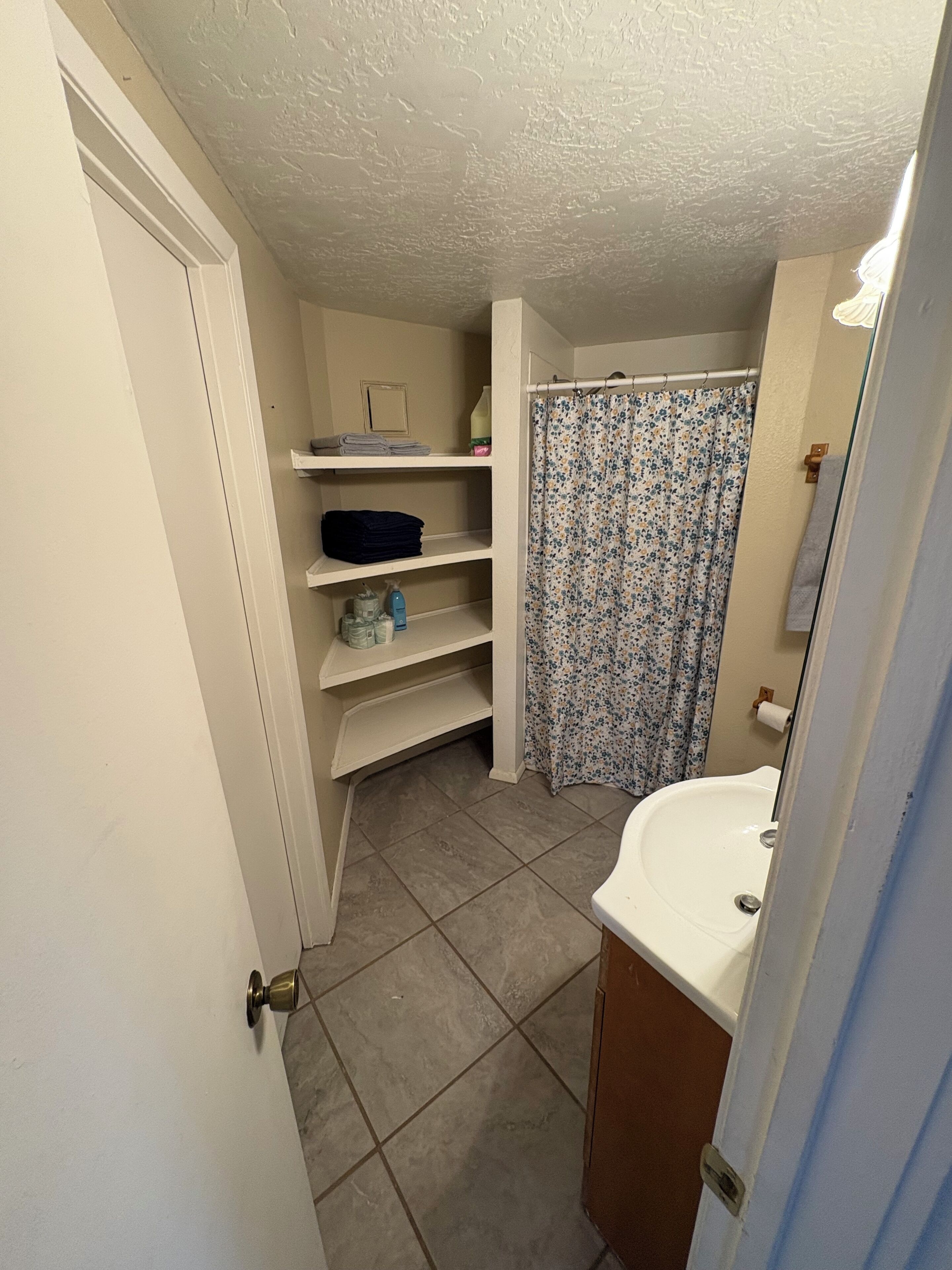 Een badkamer met een wit wastafelmeubel, een douche met een bloemen gordijn en planken met toiletartikelen.