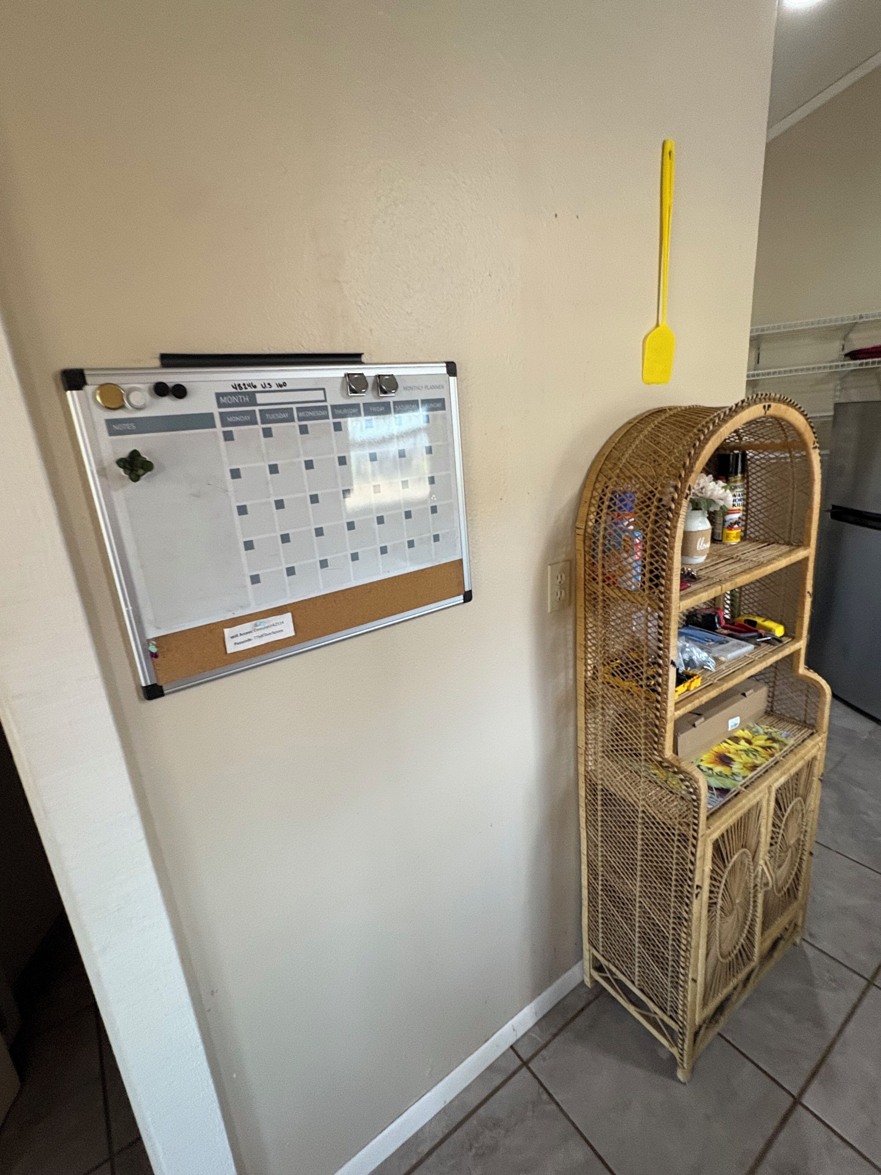 Un calendario de corcho, un estante de mimbre con varios objetos, y una espátula amarilla colgada en la pared.
