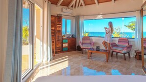 Une pièce spacieuse dotée d’une grande porte-fenêtre coulissante qui donne sur un balcon offrant une vue sur la mer. On y trouve deux chaises colorées et une petite table.