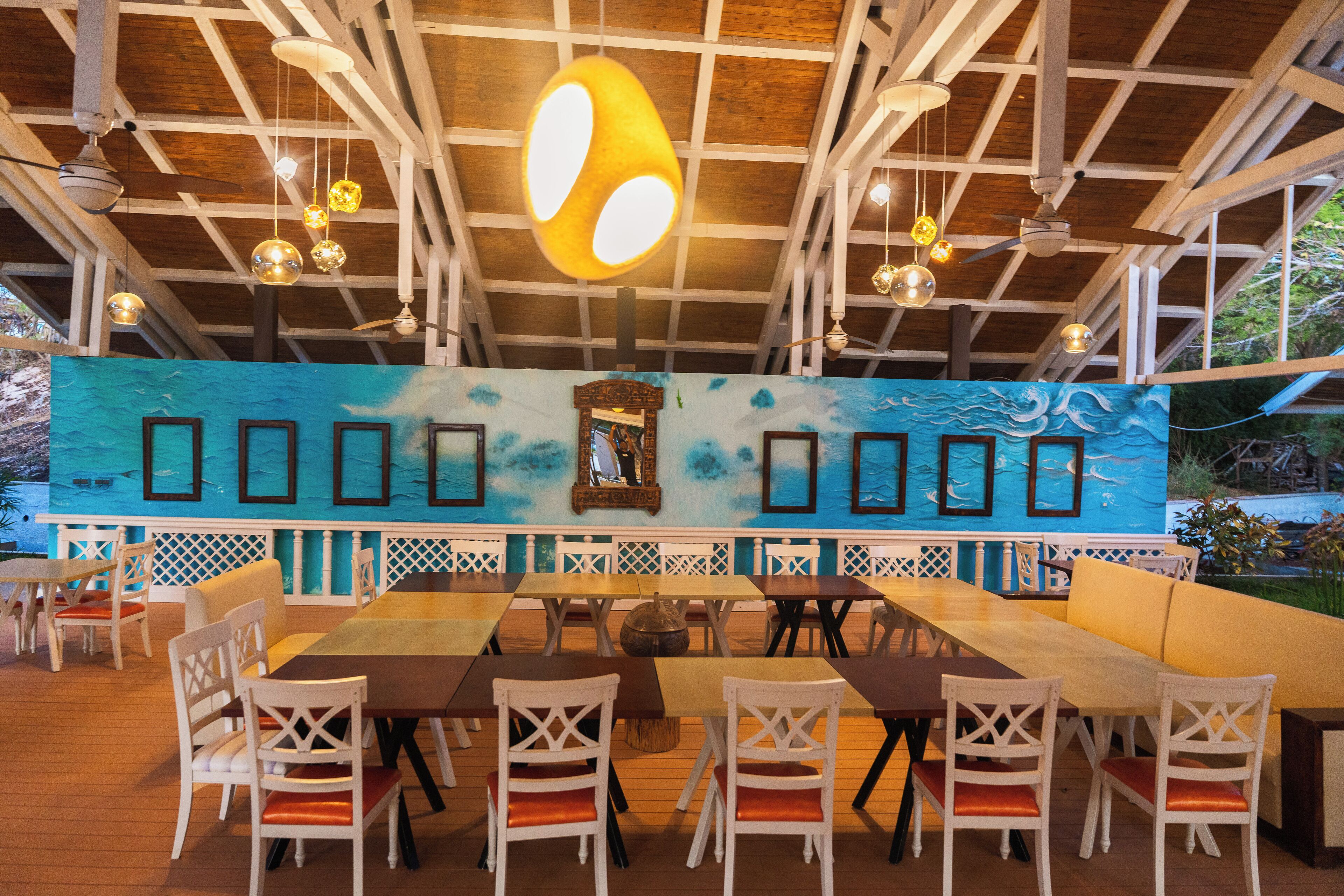 Un comedor interior con mesas de madera y sillas blancas, un mural grande en la pared y lámparas colgantes.