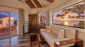 Ein gemütliches Wohnzimmer mit Holzbalkendecke, einer Couch mit Kissen, einem Couchtisch und einem großen Fenster mit Blick auf einen Sonnenuntergang über dem Wasser.