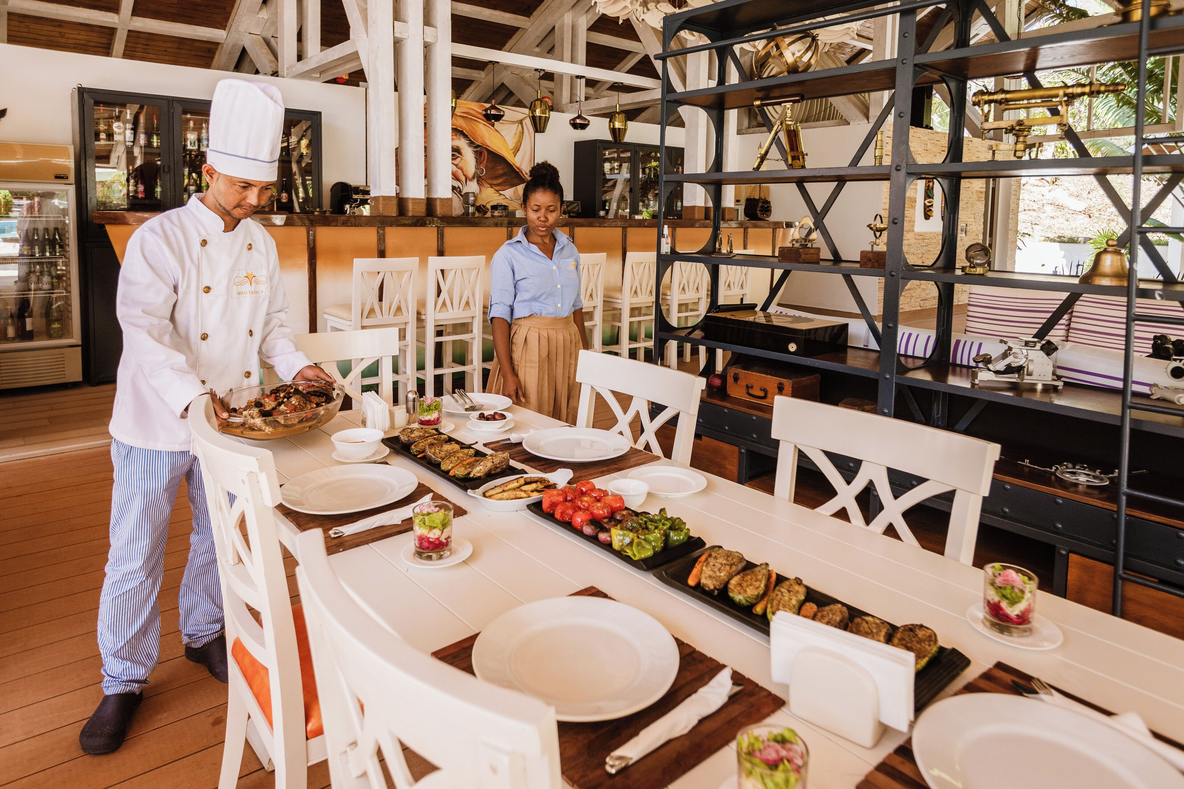 Un restaurante con una larga mesa de comedor preparada para una comida, sillas blancas y una zona de bar con estantes y objetos decorativos.