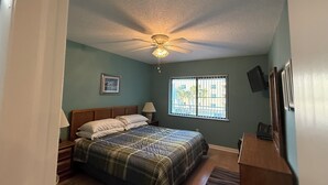 A bedroom with a bed, nightstand, lamp, ceiling fan, window, and a TV on the wall. - Unit # 321 (Cocoa Beach)