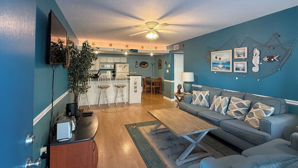 A living room with a grey sofa, a wooden coffee table, and a dining area with white chairs. - Unit # 321 (Cocoa Beach)