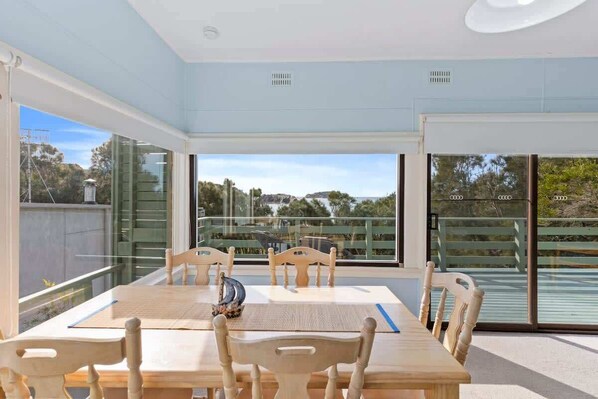 A dining area with a wooden table and chairs, large windows with a view of the outdoors, and a ceiling with recessed lighting. - Salt Shack | Beachfront Retro Escape + Pets (Guerilla Bay)