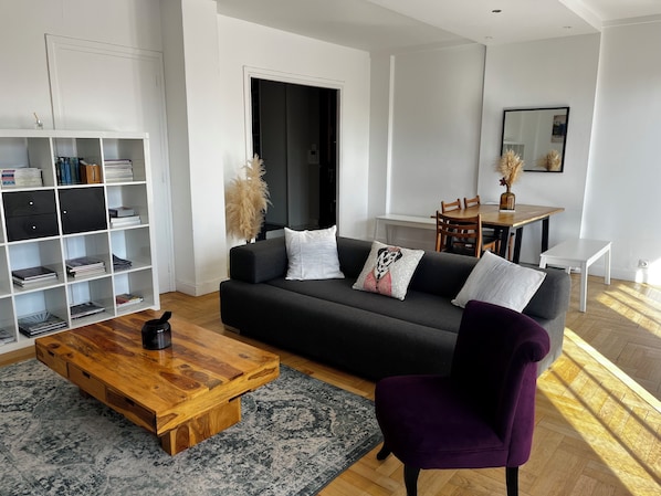 A modern living room with a dark grey sofa, a wooden coffee table, a purple armchair, a bookshelf, a dining area with a wooden table and chairs, and a large mirror on the wall.