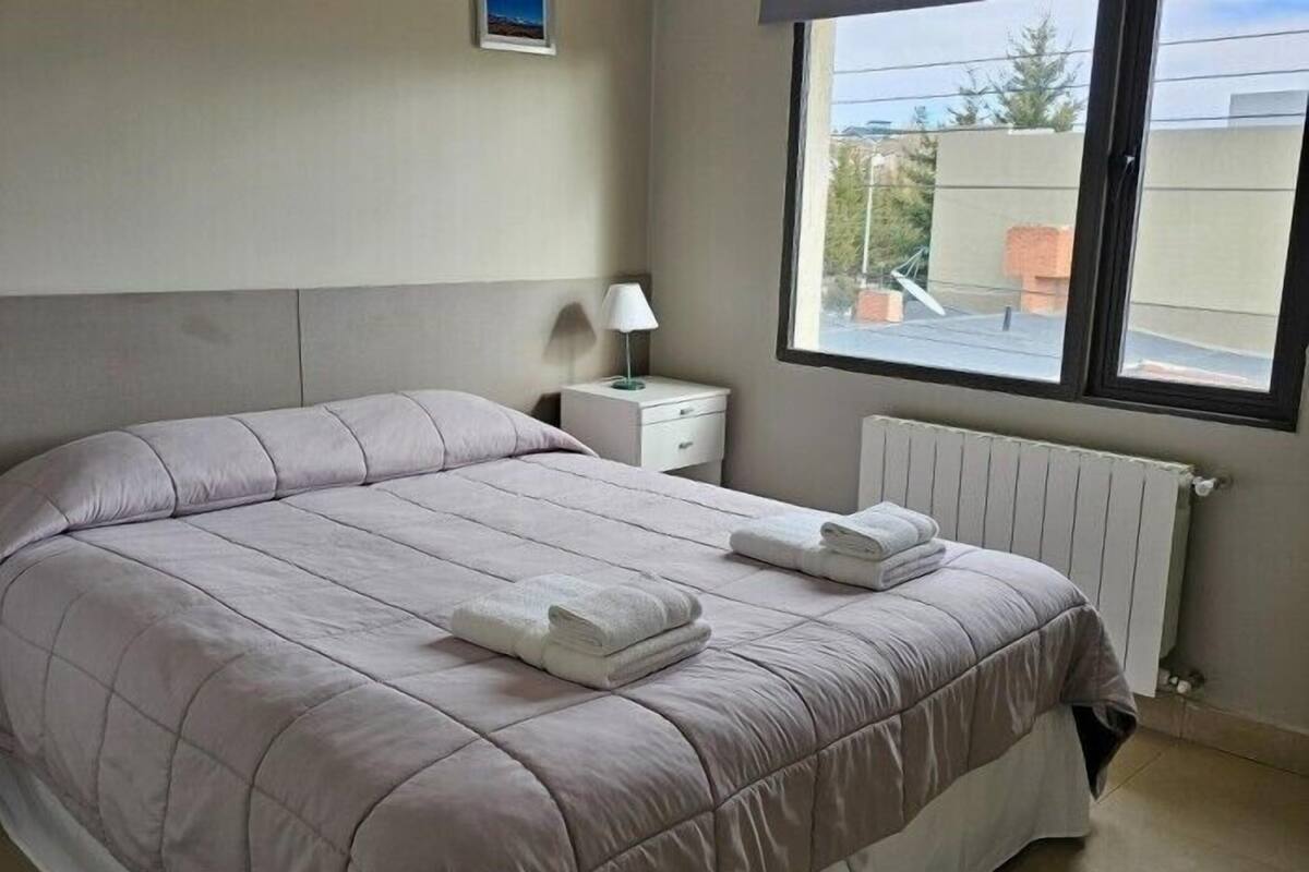 Ein Schlafzimmer mit einem Bett, einem Nachttisch, einer Lampe und einem Fenster mit Blick auf Gebäude.