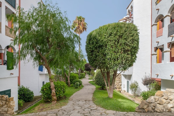 A narrow stone pathway flanked by white buildings with balconies, surrounded by greenery and a palm tree.