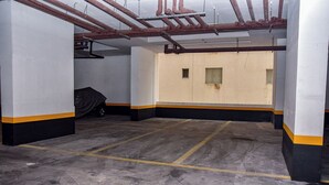 A large, empty room with a concrete floor, white walls with black and yellow trim, and visible pipes on the ceiling. - Flat com Piscina e Lazer na Praia de Pitangueiras (Guarujá)