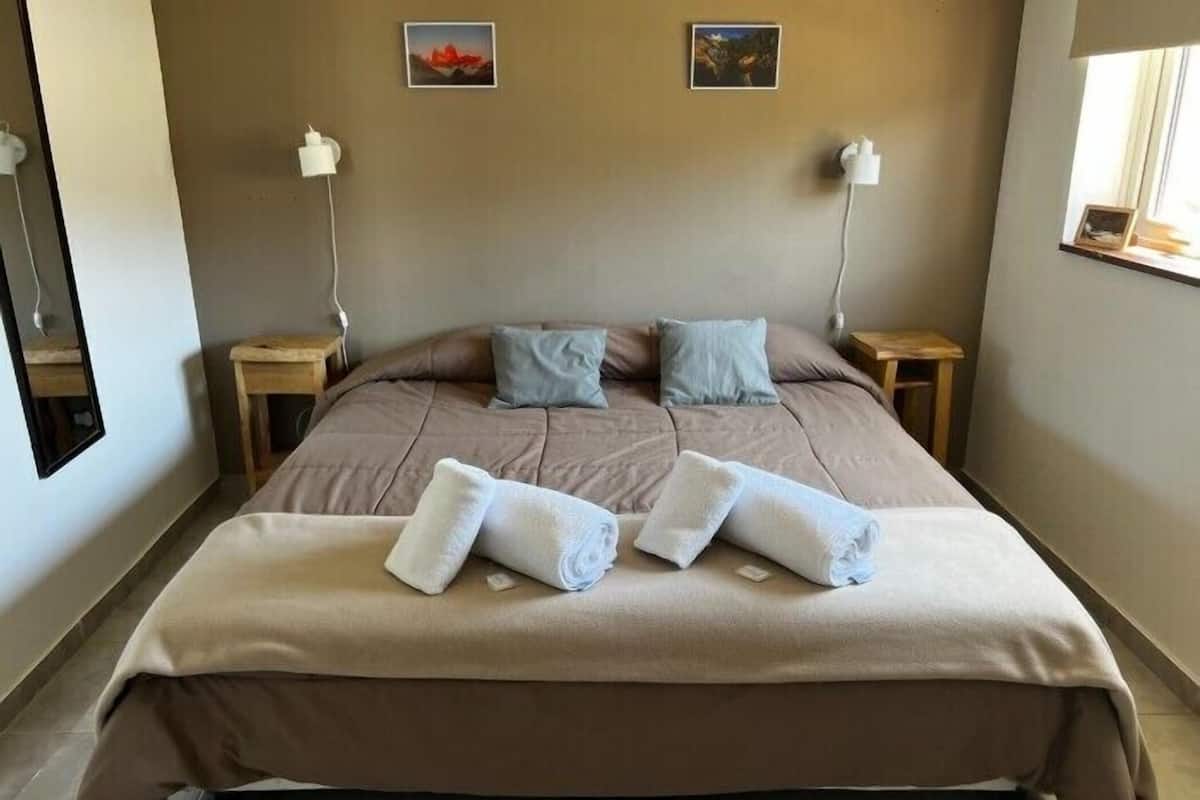 Un letto rifatto con cura, con copriletto color beige e due cuscini, un piccolo comodino in legno con una lampada e un quadro appeso al muro.