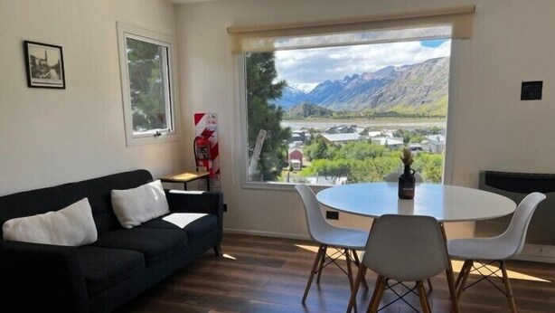 Ein Wohnzimmer mit einer schwarzen Couch, weißen Sesseln, einem kleinen Tisch und einem großen Fenster mit Blick auf die Berge.