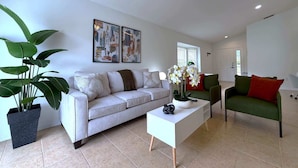 Un salon moderne avec un canapé, une table basse et un fauteuil vert. Deux œuvres d’art encadrées ornent le mur, ainsi qu’une plante en pot et un ventilateur de plafond.