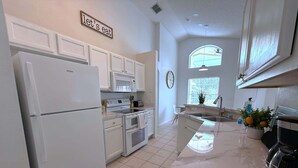 Une cuisine moderne avec des meubles blancs, un four encastré, un micro-ondes et un réfrigérateur. Il y a une fenêtre donnant sur l’extérieur et une horloge au mur.