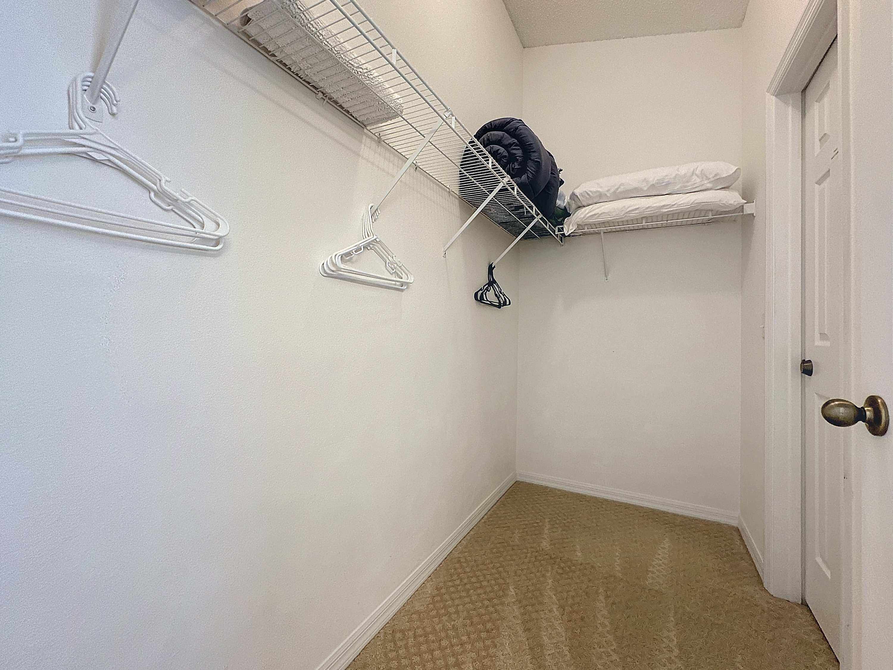 A small, narrow hallway with a white door, a white wall, and a beige carpet. There is a white wire shelf with clothes and a black hanger on it.