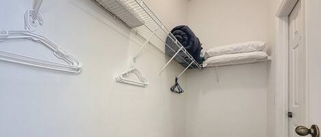 A small, narrow hallway with a white door, a white wall, and a beige carpet. There is a white wire shelf with clothes and a black hanger on it.