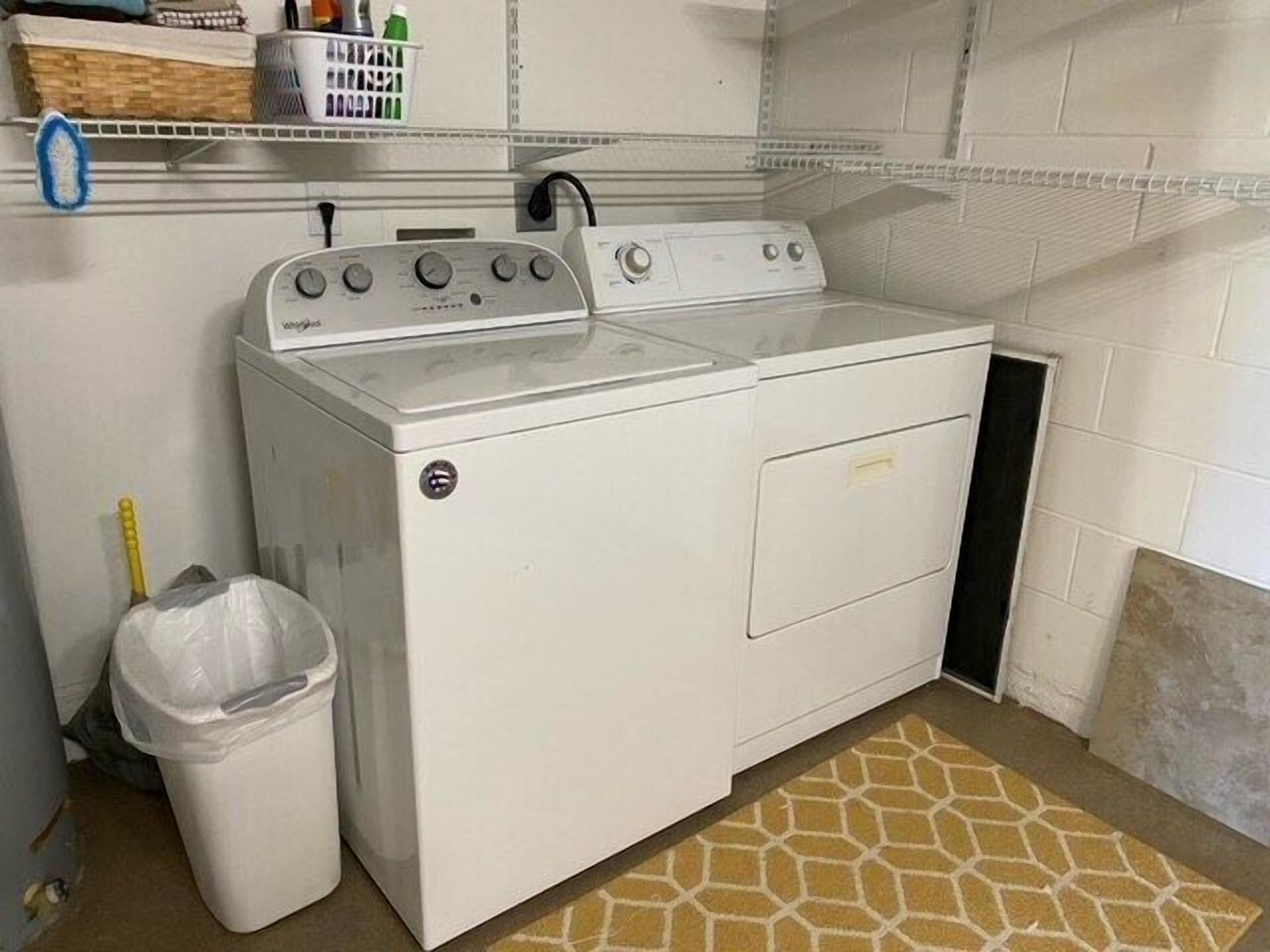 A compact, white front-loading washer and dryer unit in a laundry room with a tiled backsplash, a small trash bin, and a patterned rug.