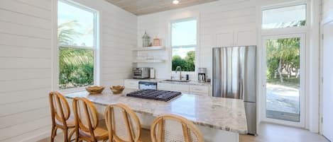 A modern kitchen with white cabinetry, a large island with bar stools, a stainless steel refrigerator, and a view of greenery outside.