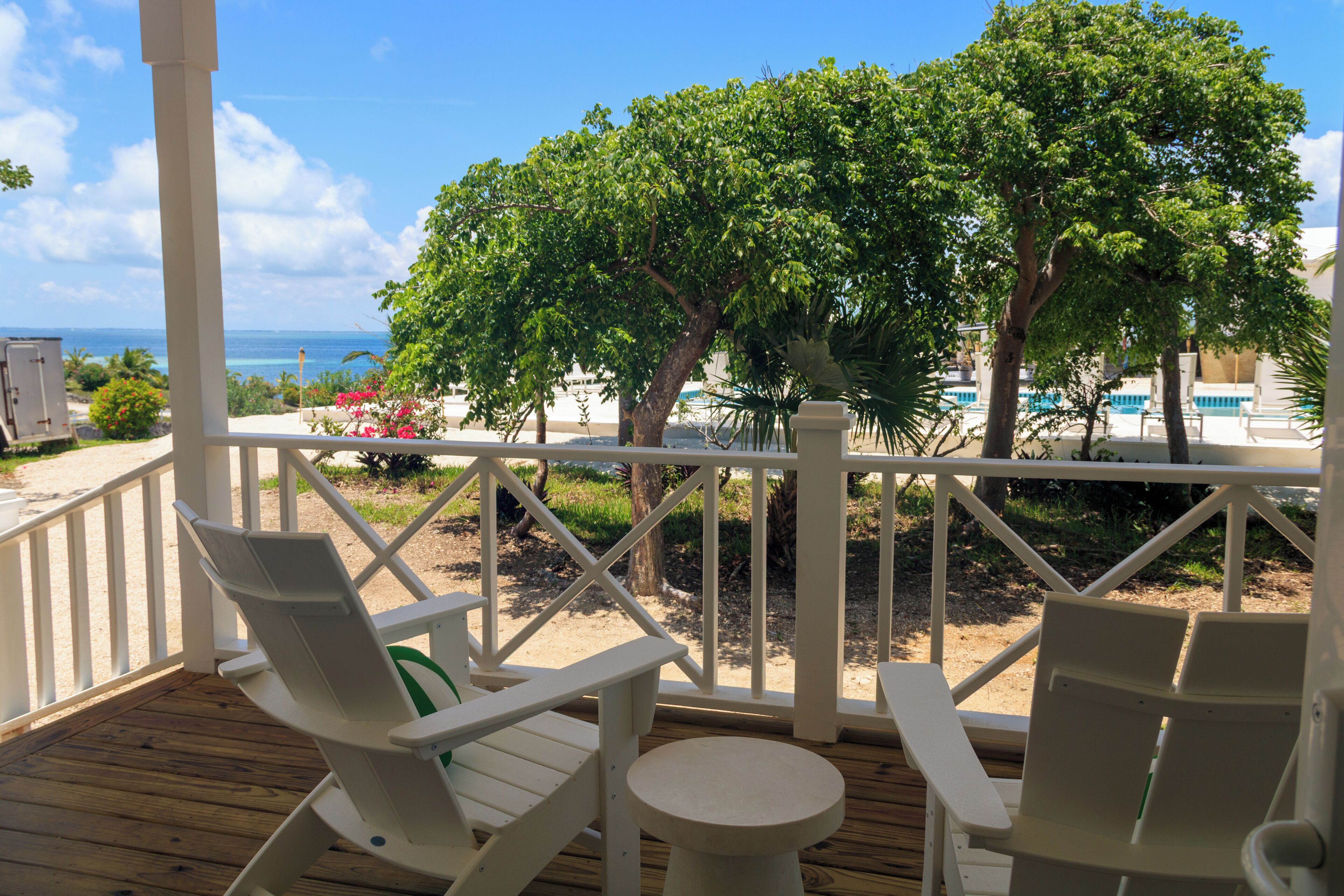 Um deck de madeira com cadeiras brancas e uma pequena mesa, com vista para uma praia com piscina e árvores.