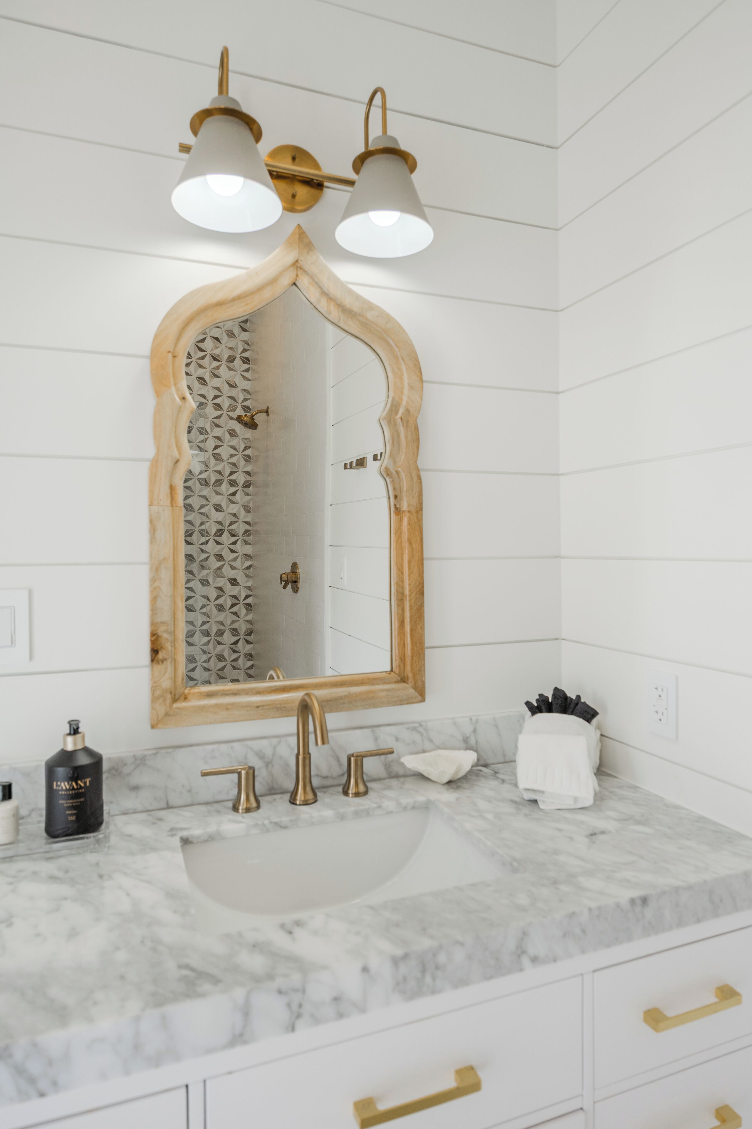 Un baño con lavamanos de mármol, accesorios dorados, espejo con marco de madera y pared de ducha con diseño.