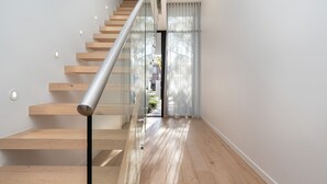Un escalier moderne avec des marches en bois et une rampe en verre, menant à une porte qui offre une vue sur la verdure à l’extérieur.