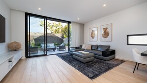 Un salon moderne avec un canapé gris, un tapis noir et une grande porte-fenêtre coulissante donnant sur une terrasse. Deux œuvres d’art encadrées ornent le mur, et un téléviseur à écran plat y est fixé.