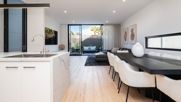 Une cuisine moderne dotée d’un grand îlot central, d’un coin repas avec une table sombre et des chaises blanches, et offrant une vue sur un jardin grâce à de grandes portes coulissantes.