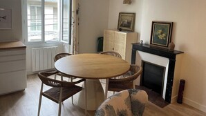 A dining area with a round wooden table and wicker chairs, a white cabinet, and a fireplace with a framed picture above it. - Renovated apartment - near beach (Dinard)