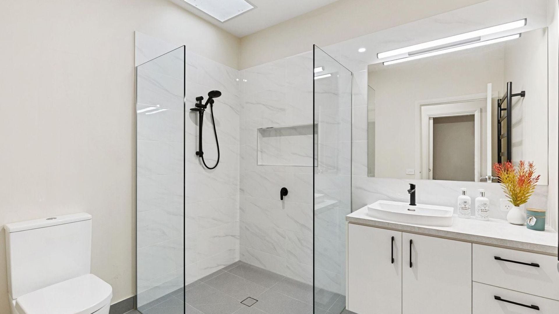 Une salle de bain moderne avec une douche à parois de verre, des toilettes blanches, un lavabo avec un miroir et un vase rempli de fleurs.