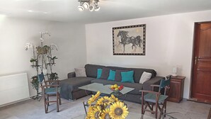 A living room with a sectional sofa, a glass coffee table with sunflowers, a metal chair, a potted plant, and a painting on the wall.