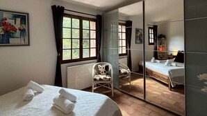 A hotel room with a bed, a chair, a window with curtains, a painting on the wall, and a mirror reflecting the room.