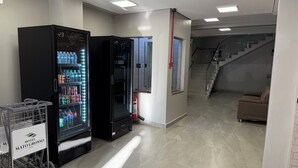 A hotel lobby with two large black refrigerators stocked with beverages, a fire extinguisher, a staircase, and a brown sofa. - Hotel Mato Grosso Inn (Cuiabá)