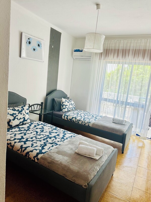 A hotel room with two beds, a table, a chair, a ceiling fan, and a window with curtains.