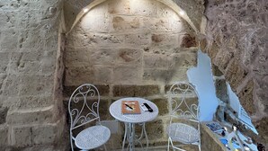 Une pièce aux murs de pierre, avec deux tables et chaises en métal blanc, une petite table sur laquelle sont disposés divers objets, et un luminaire fixé au mur.