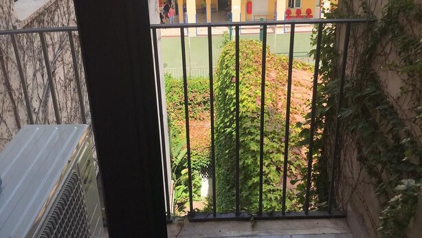 Een balkon met een metalen hekwerk, een uitzicht op een gebouw met meerdere ramen en balkons, en wat groen op de achtergrond.