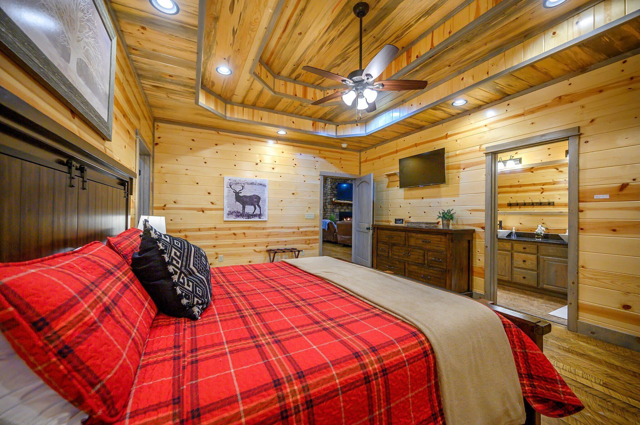 Een slaapkamer in een houten chalet met een bed, een plafondventilator, een wandtelevisie en een houten deur naar een badkamer.
