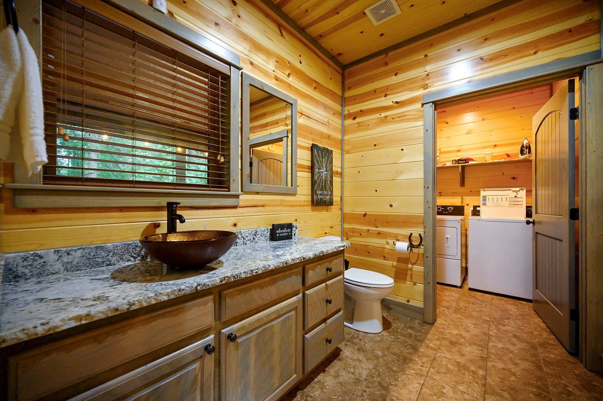 Une salle de bain rustique, avec des murs en bois, un comptoir en granit, un lavabo sur pied, des toilettes et un petit coin buanderie.