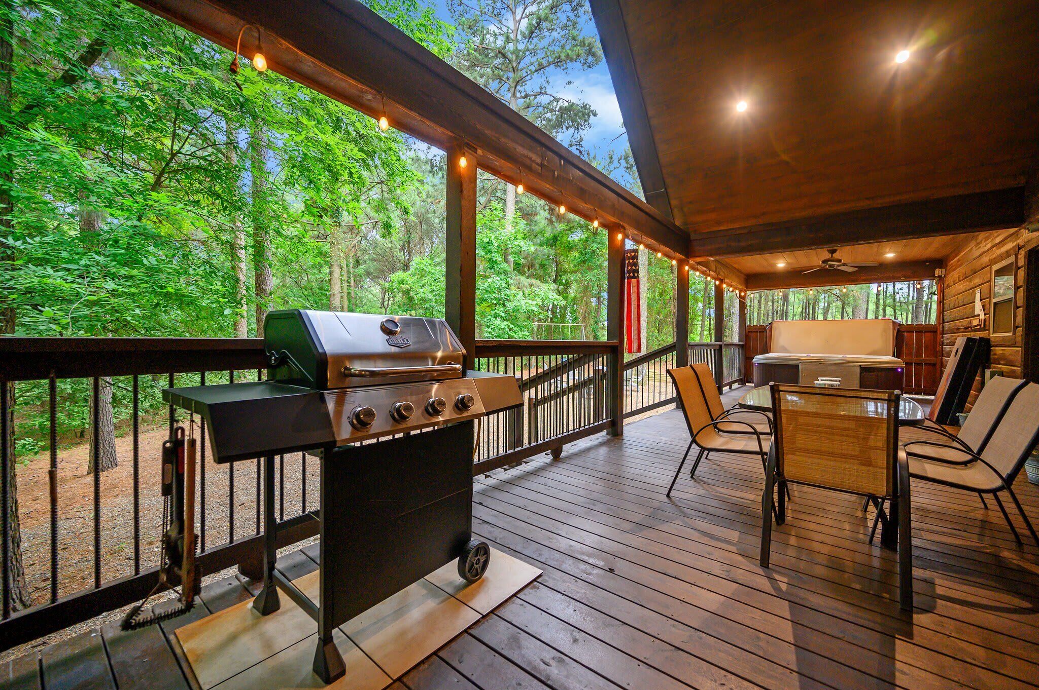 Een terras buiten met een barbecue, stoelen en een tafel, omringd door een houten balustrade en met uitzicht op een bos.