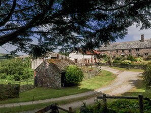 A rural setting with a stone building, a thatched-roof structure, and a paved path leading to a larger stone building with multiple windows. - Lane End (South Molton)