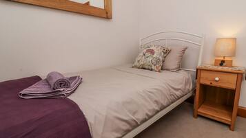 A single bed with a beige bedspread and two pillows, one with a floral pattern. A purple towel is folded on the bed. A wooden nightstand with a lamp and a small drawer is beside the bed.