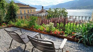 A terrace with two metal chairs, a garden with various plants and flowers, and a view of a lake and mountains. - Elegant Apartment in the Heart of Bellagio (Bellagio)