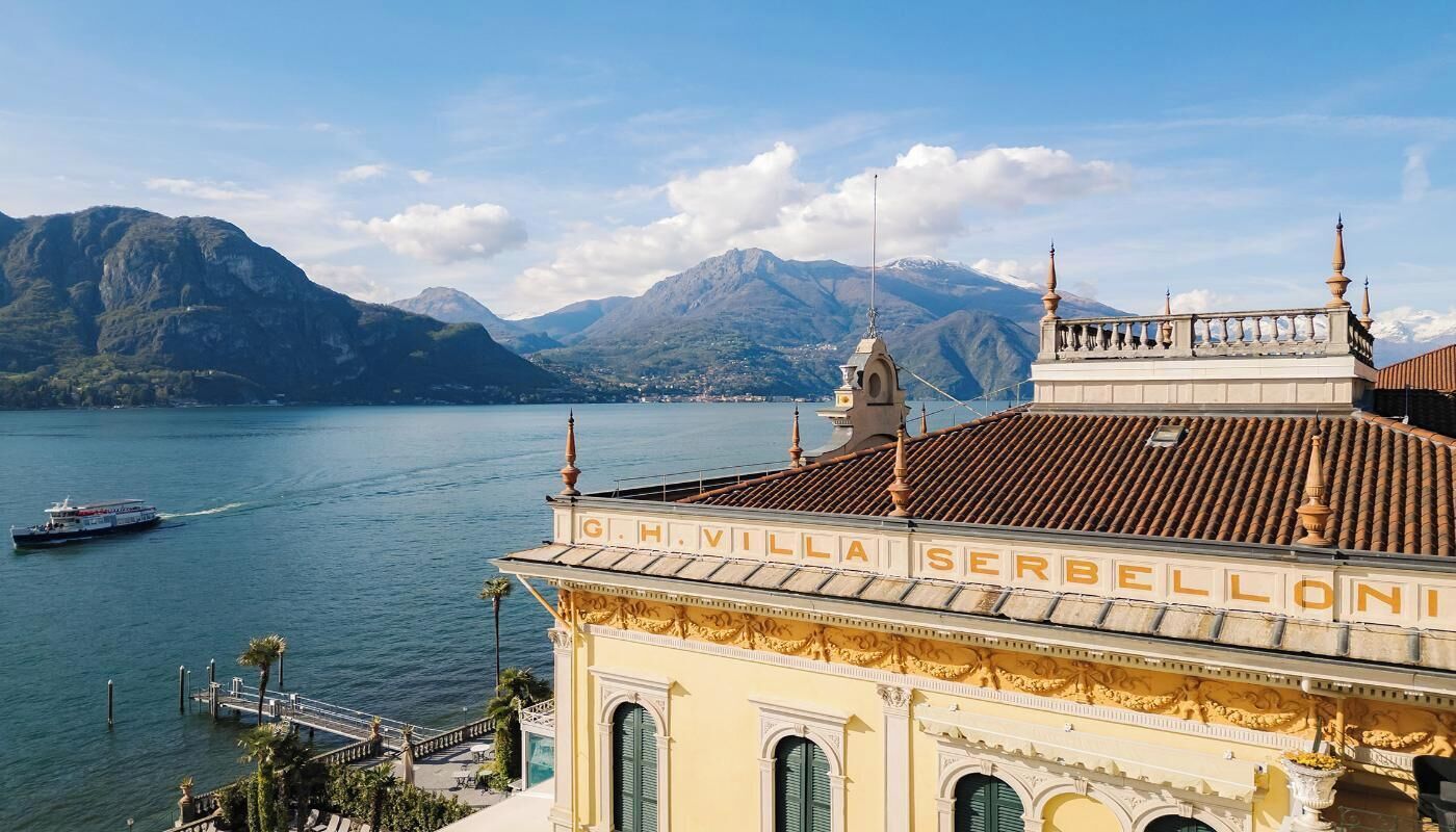 A building with the sign "G.H. Villai Serbelloni" overlooking a lake with mountains in the background.