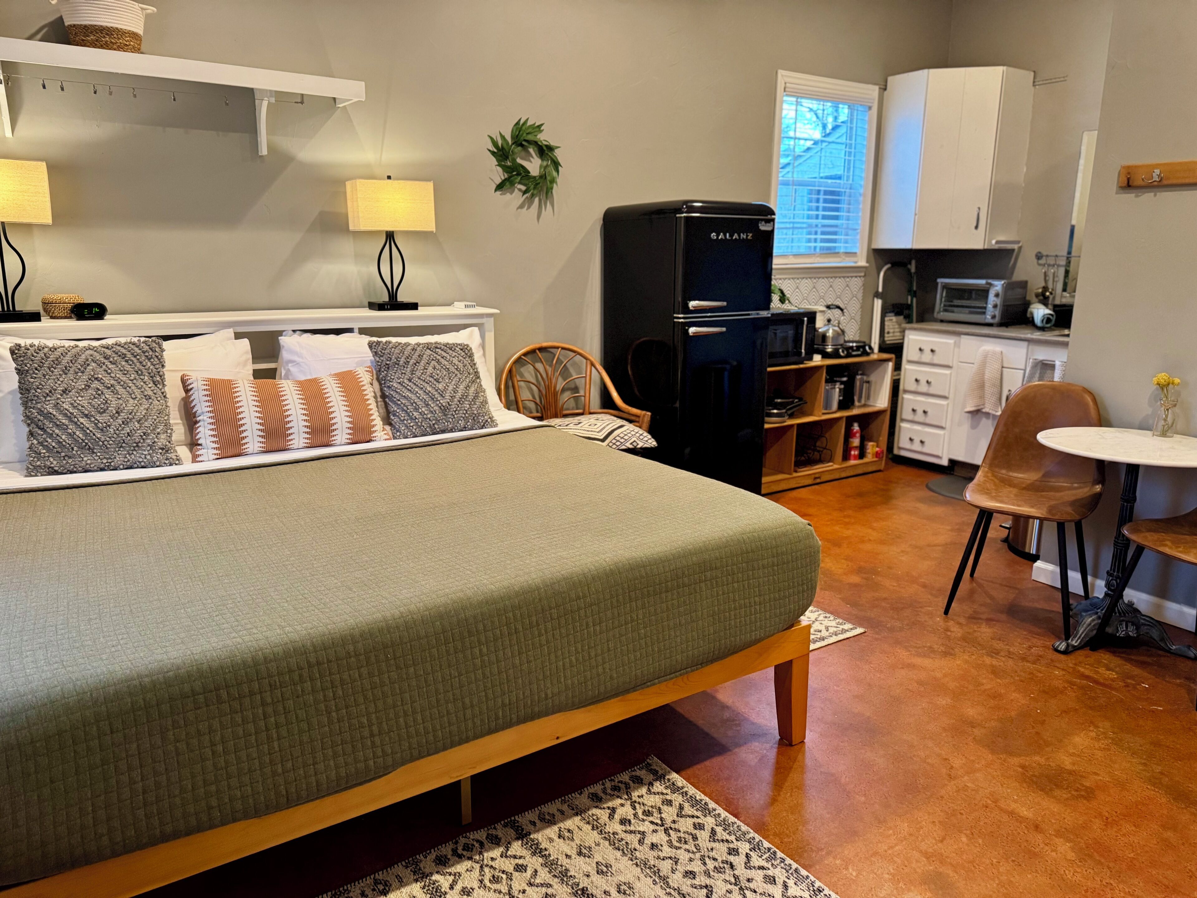 A cozy bedroom with a bed, bedside lamps, a small desk, and a kitchenette.