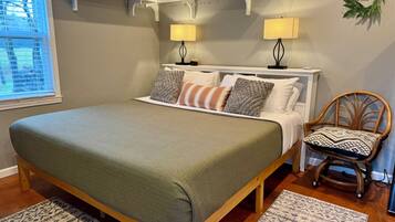 A bedroom with a large bed, two bedside lamps, a wooden chair, a shelf with decorative items, and a window with blinds.