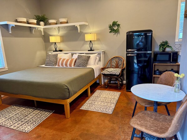 Un dormitorio compacto con cama, lámparas de noche, una silla, una mesita, un refrigerador y un microondas.