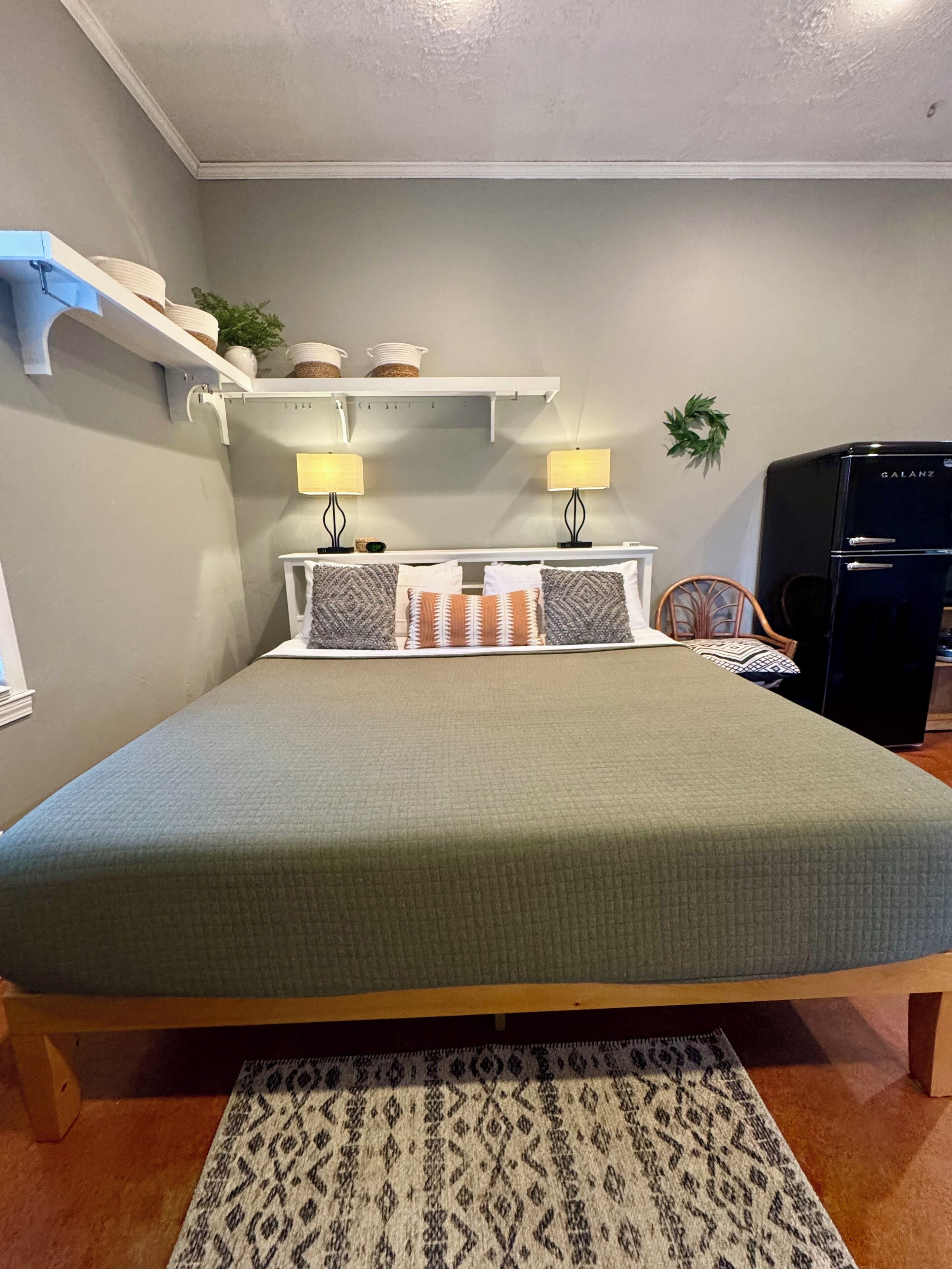 A bedroom with a bed, bedside lamps, a shelf with decorative items, a small plant, and a refrigerator.