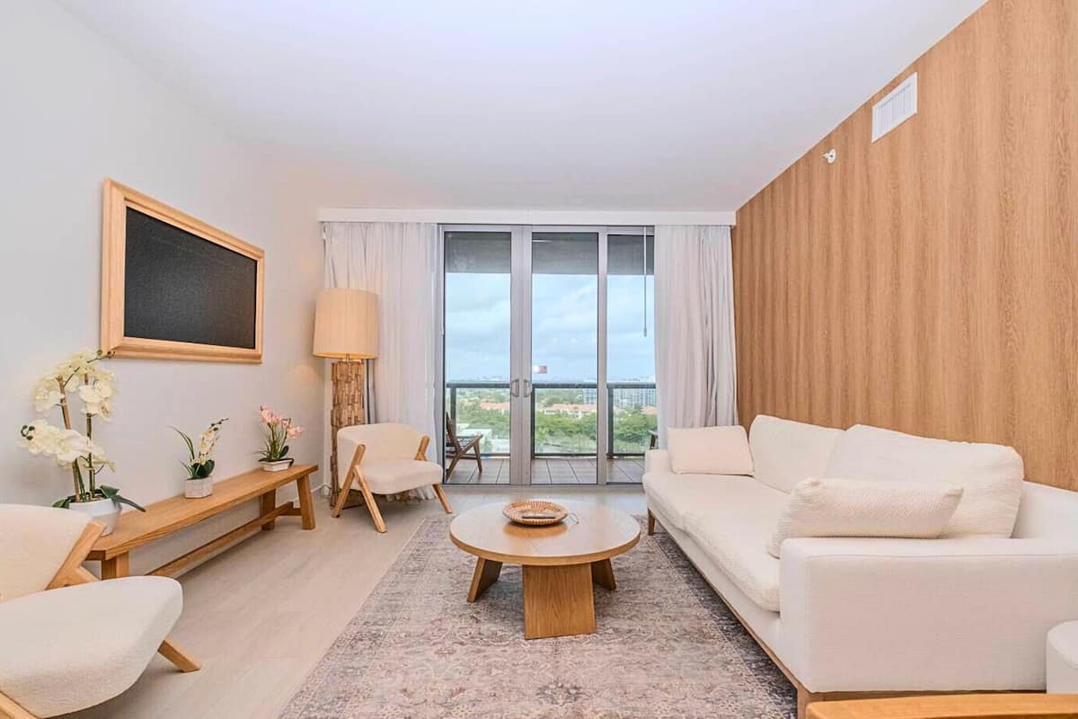 Un salon moderne avec un canapé blanc, une table basse ronde et un banc en bois. Une grande fenêtre avec des rideaux, une lampe et un tableau encadré ornent le mur.
