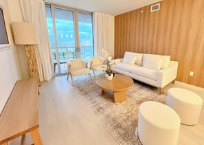 Una sala de estar moderna con un sofá blanco, dos sillones, una mesita de centro y vistas a un paisaje urbano a través de grandes ventanales.